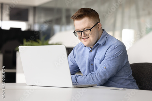 Smiling professional male office manager with Down syndrome working on laptop, review tasks to-do, engaged in administrative remote workflow seated at desk in contemporary workspace. Inclusivity, tech