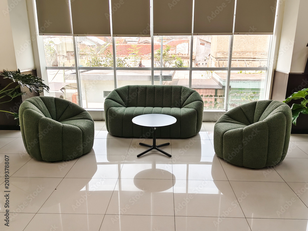 Fototapeta premium Modern green sofa set sits beneath large windows. Circular table rests at center, inviting conversation. Bright tiles reflect light, enhancing open space. Potted plants add natural touch