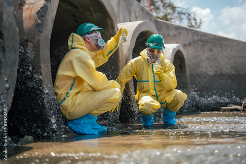 Environmental inspectors analyze contaminated water samples while working beside industrial sewage pipes. Protective equipment evaluate pollution risks and wastewater treatment.