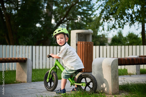 Toddler riding balance bike in park, wearing helmet and looking back. Active outdoor childhood, learning coordination and safe cycling habits in sunny urban environment