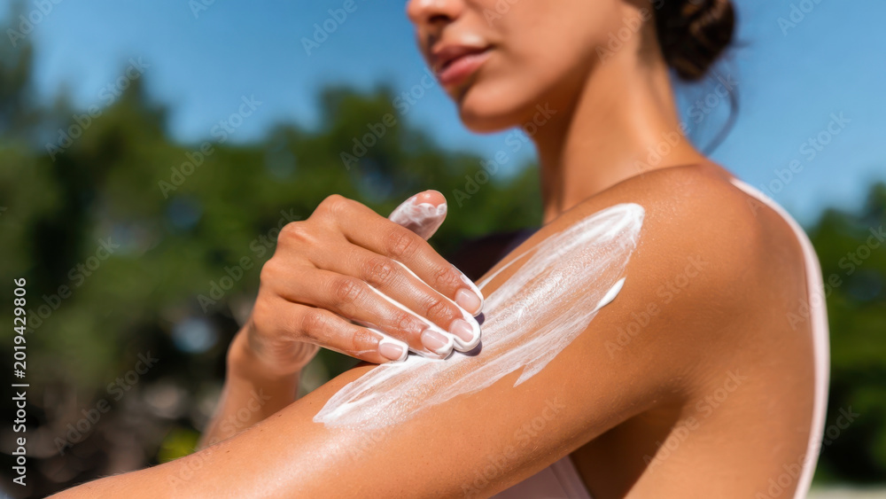 Fototapeta premium Young woman applying sunscreen on arm in sunny outdoor setting with blurred trees