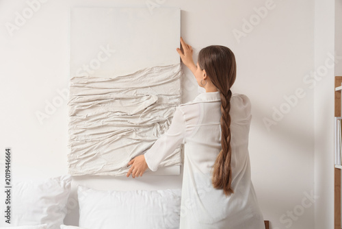 Quadro su tela Young woman hanging 3D painting on light wall in bedroom, back view
