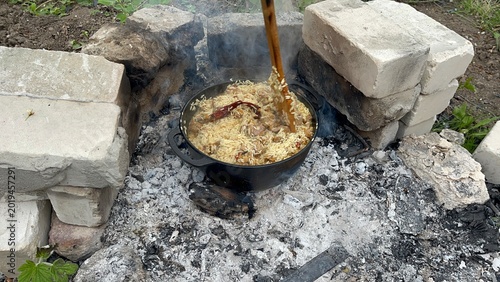Fotografie Traditional pilaf cooking in a large cast-iron cauldron over an open fire supported by bricks outdoors