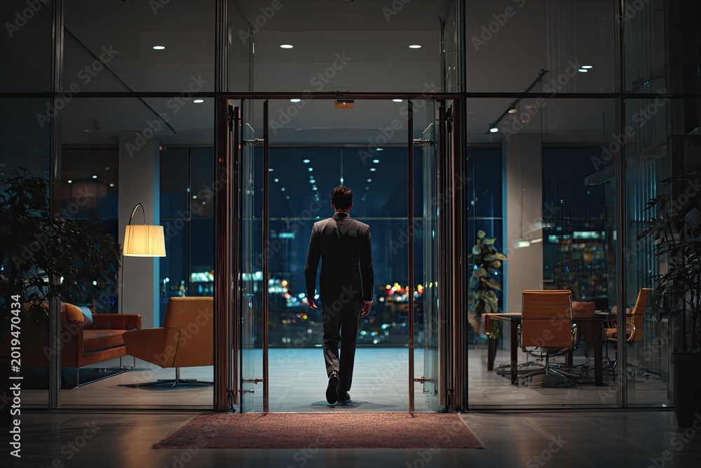 Fototapeta premium Man in suit walks through glass doors at night into modern office
