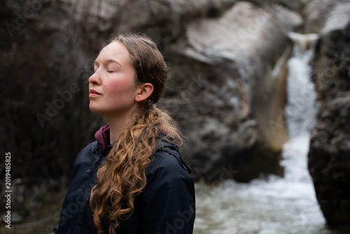 A peaceful female practices breathing outdoors near a waterfall. A young woman breathes in the fresh air of nature, restoring her mental health.
