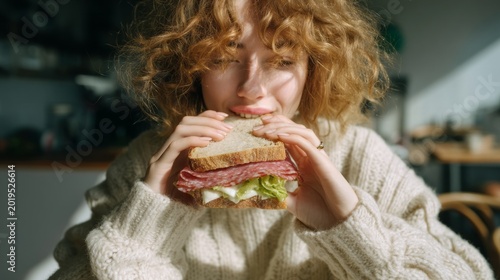 Bright sunlight bathes a young individual with curly hair as they prepare to take a big bite of a delicious sandwich. The cozy indoor atmosphere invites joy and comfort