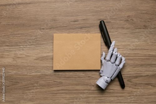 High angle view of a white mechanical robotic hand holding a black pen next to a blank brown card on a wooden surface.