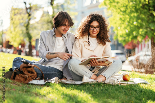 Young couple sitting on grass in a park, engaged in reading a book together, surrounded by trees and a sunny outdoor environment