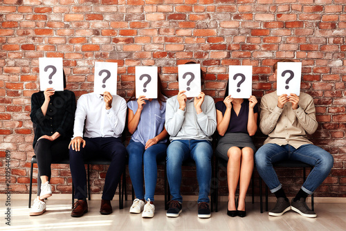 Group of diverse people sitting in a row holding question mark signs over their faces expressionless