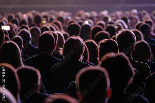 Large audience watching live performance in dimly lit venue, backs of heads visible, phones recording, atmosphere of anticipation and focus.