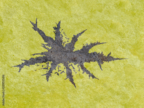 Abstract illustration featuring a dark, jagged ink-like splash or splatter centered on a vibrant, textured lime green and yellow background with a rough, paper-like surface.

