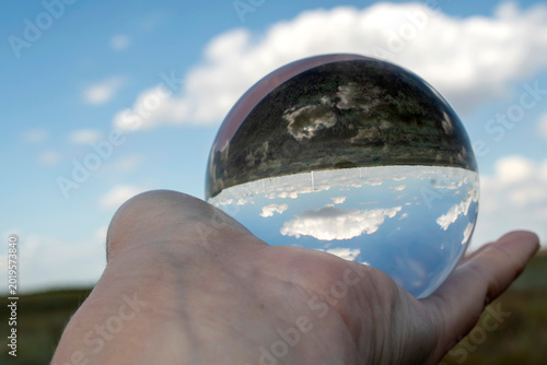 Creative photography of blue sky and white clouds refracted inside a glass sphere held by a human hand. Optical illusion and clean environment concept.