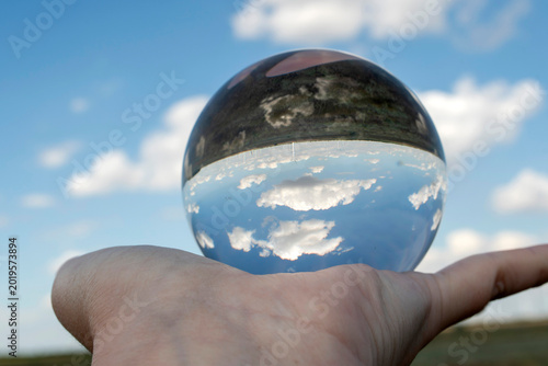 Creative photography of blue sky and white clouds refracted inside a glass sphere held by a human hand. Optical illusion and clean environment concept.