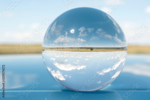 Minimalist concept with a glass sphere reflecting blue sky and clouds, resting on a sleek, glossy blue metallic surface. Futuristic abstract background.