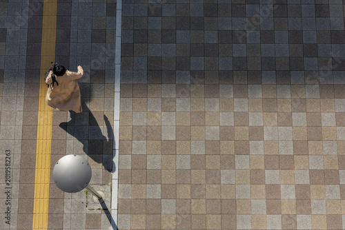 Cuadro en lienzo Commuter walking with authority in early morning sunlight, coat hem swaying with rhythm as her shadow stretches long across the tiles of an empty urban plaza with lone streetlamp