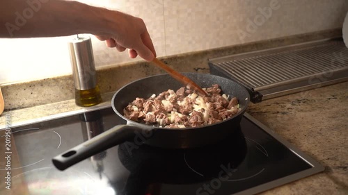 Male hand stirring diced meat and onion with a wooden spoon in a pan on an induction stove