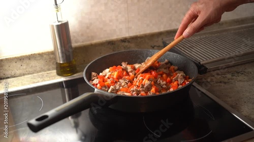 Close-up of a frying pan with beef, onion, and diced red sweet pepper being cooked on a stove in a home kitchen.