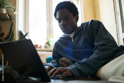 Fotografie African American teenage boy using laptop while lying on sofa at home, studying online and browsing digital content