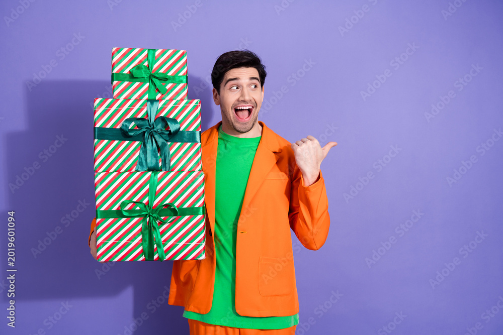 Naklejka premium Excited young man in orange jacket holding stacked Christmas gifts and pointing with joy at colorful holiday presents