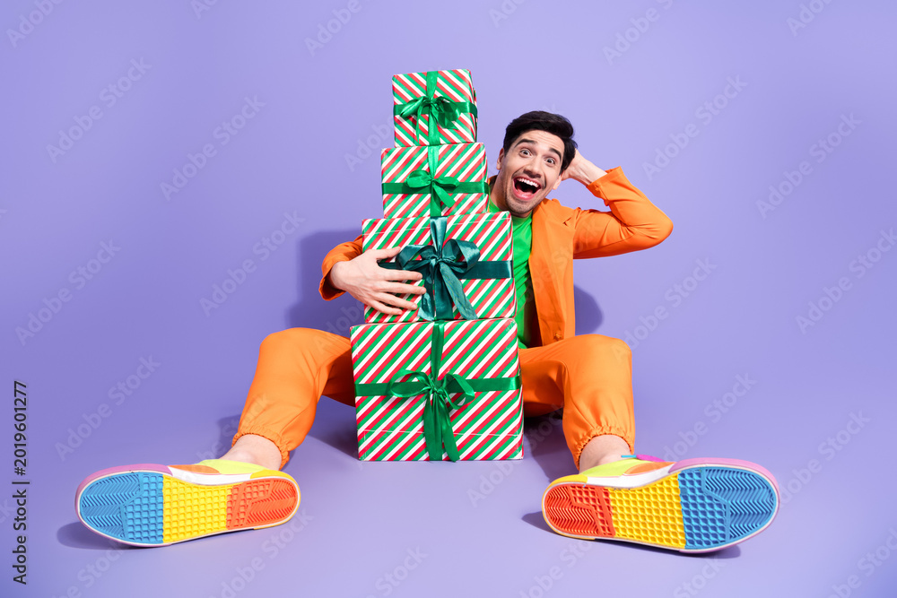 Obraz premium Happy man in orange outfit sitting with stack of wrapped Christmas gifts celebrating colorful holiday excitement