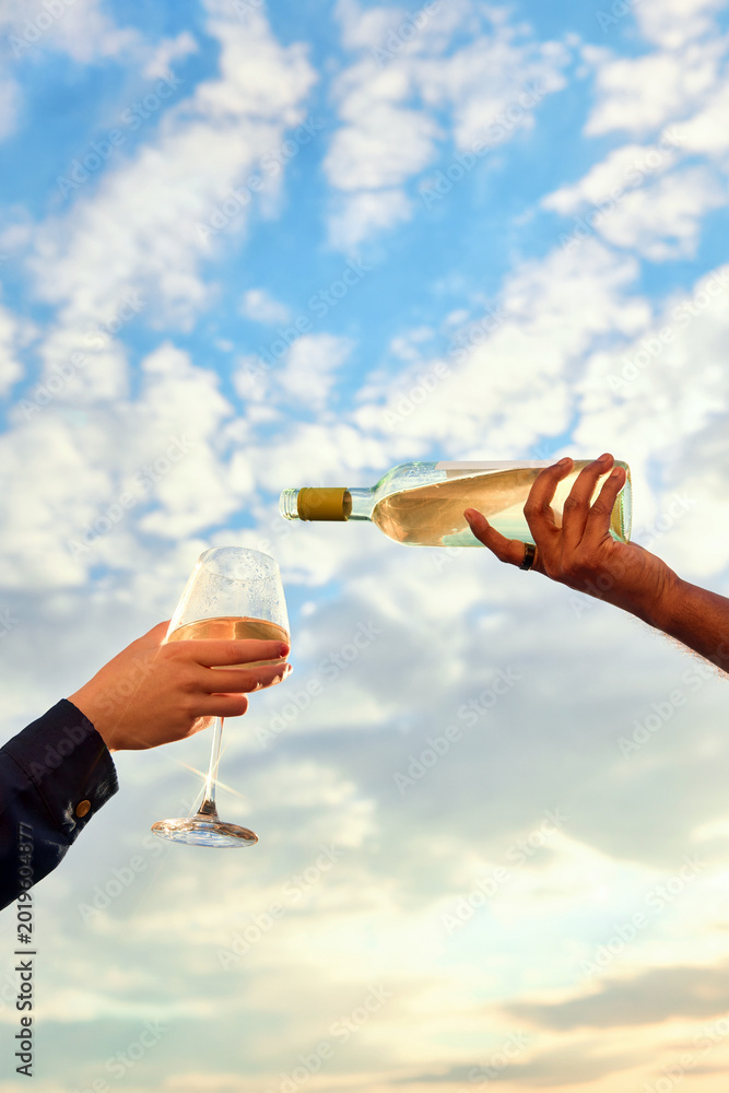 Fototapeta premium Hand pouring white wine into glass with cloudy blue sky background. Concept of vineyard tourism, wine tasting, romantic getaway, premium beverage branding and outdoor dining.