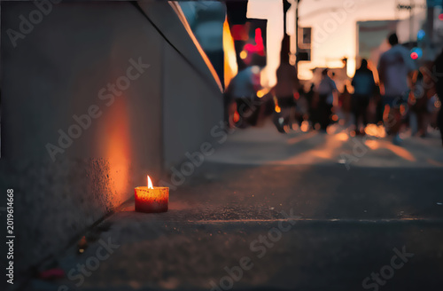 Lone candle glows on sidewalk as blurred figures walk past at dusk