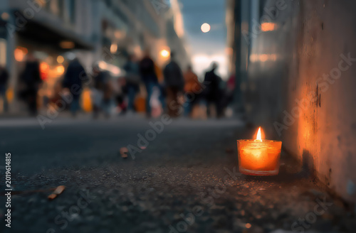 A solitary lit candle glows warmly on a street, illuminating a blurred background of walking figures