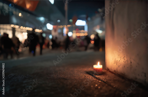 A solitary candle glows on a snowy street at dusk, illuminating the blurred figures of a crowd