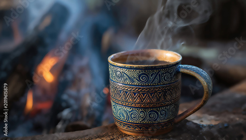 Steaming mug of dark beverage sits by a glowing campfire at dusk