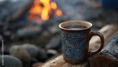 Steaming mug of dark beverage on driftwood by a campfire