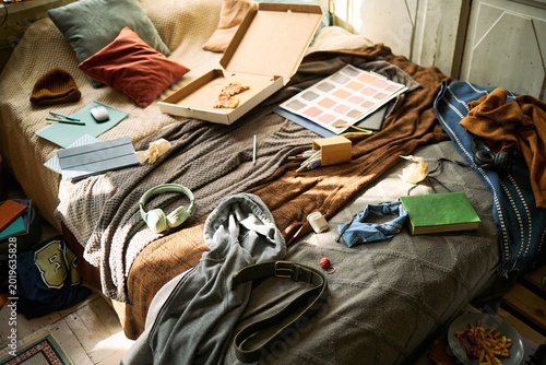 Fotografie Wide angle shot of teenager bedroom showing bed covered with school supplies, pizza box, headphones, clothing, signaling messy student routine