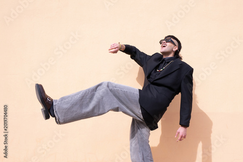A young man laughing with one leg raised in a dynamic pose in front of a peach-colored outdoor wall