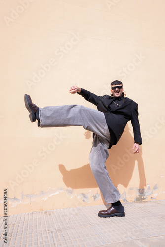 A young man laughing with one leg raised in a dynamic pose in front of a peach-colored outdoor wall