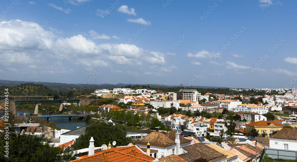 Fototapeta premium Tavira shows white houses and narrow streets from above, highlighting the tow's layout, greenery, and surrounding hills on a clear day in Algarve, Portugal