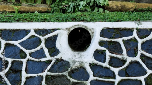 Foto A circular drainage pipe outlet integrated into a decorative stone and mortar retaining wall, surrounded by lush green garden foliage and groundcover plants in a landscaped outdoor environment