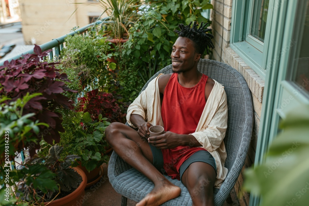 Naklejka premium Young man enjoying morning coffee on a plant-filled balcony chair for social media background and lifestyle banner
