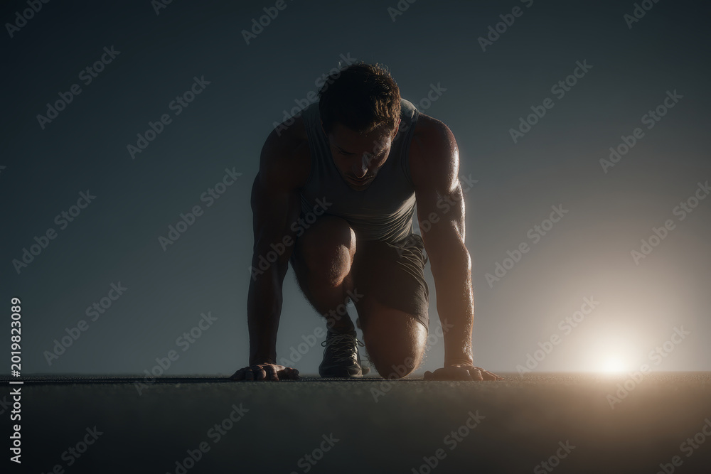 Obraz premium Athletic man in sportswear prepares to sprint at dawn, crouched in a starting position on an outdoor track. Focused runner readies himself for an intense early morning workout session