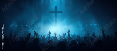 Brightly illuminated christian cross centered over crowd with raised hands during worship under blue light