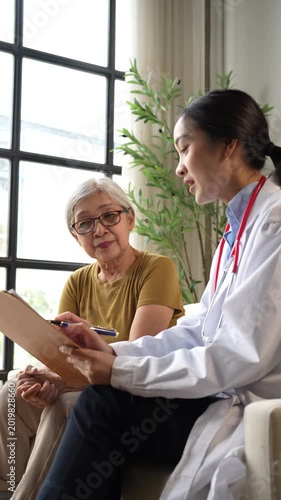 Asian female doctor explaining medical test results on a clipboard to an elderly patient during a home visit, providing geriatric care and health consultation