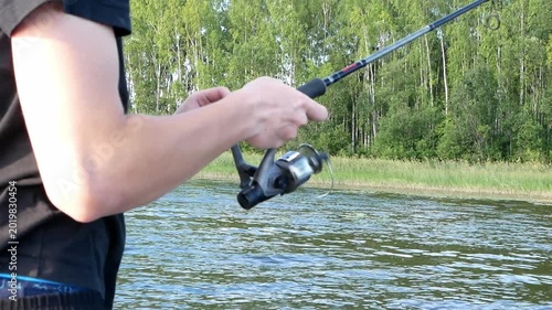 Young male fisherman catching fish with a spinning rod on a calm lake during a sunny summer day.