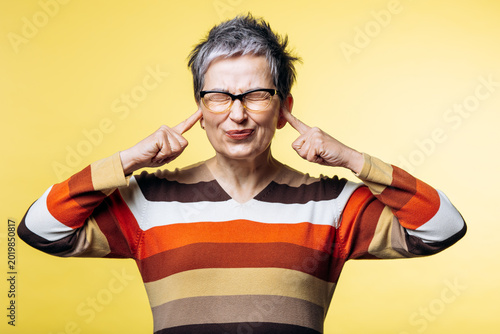 Mature woman covering her ears with fingers, expressing frustration with loud sound, standing on a yellow background