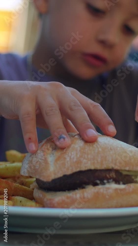 Boy grabbing hamburger with both hands during outdoor family meal capturing excitement, appetite and authentic childhood enjoyment at restaurant