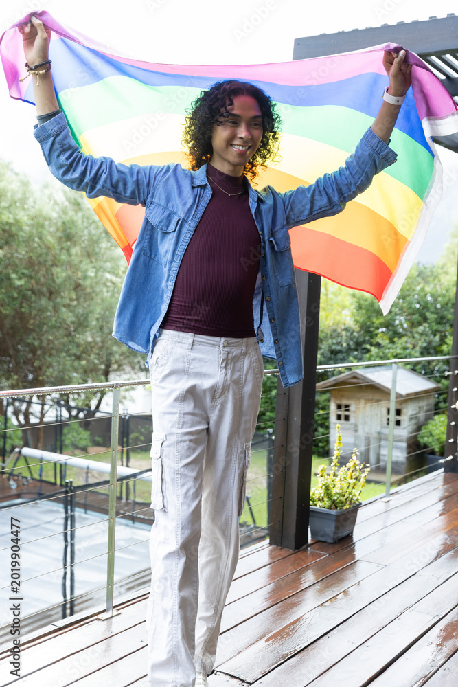 Fototapeta premium Non-binary person standing on wet deck holding rainbow pride flag, wearing denim overshirt