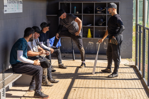 Photos Five male players gathering on left bench in baseball dugout, holding bat and gl