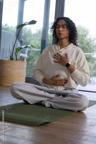 Non-binary person meditating with eyes closed on green yoga mat by glass doors, smartwatch visible