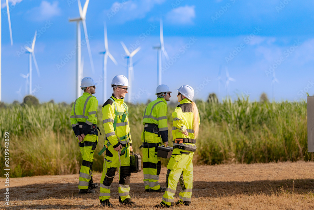 Naklejka premium A team of engineers in high visibility safety gear stand together at a wind farm, conducting inspections and discussing renewable energy projects. Wind turbines surround them under a clear sky.