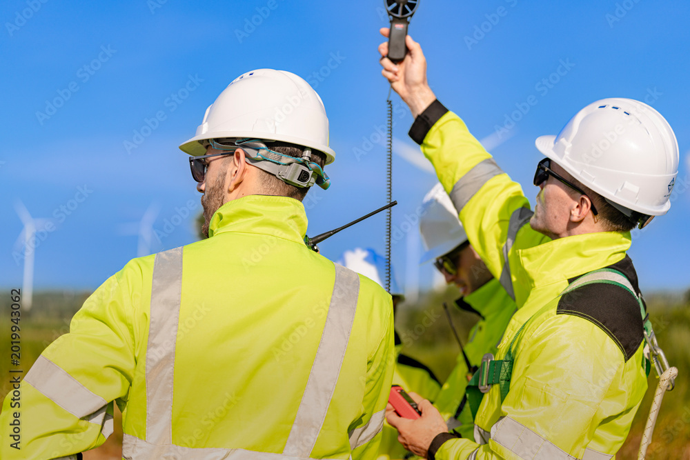 Fototapeta premium A team of engineers in high visibility safety gear stand together at a wind farm, conducting inspections and discussing renewable energy projects. Wind turbines surround them under a clear sky.