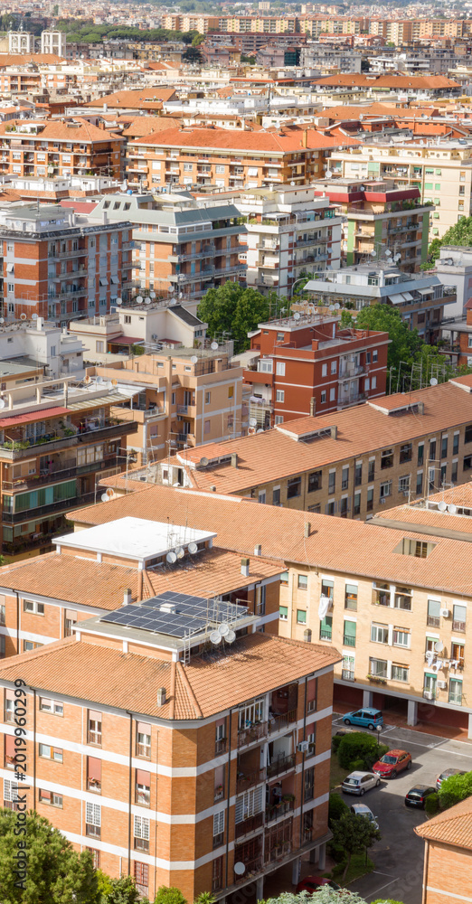 Obraz premium Aerial and vertical view of a dense urban residential area featuring numerous multi-story apartment buildings with terracotta roofs, solar panels, green trees, and parked cars in a sunny morning.