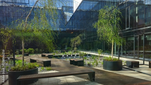 Obraz na plátne Modern urban courtyard surrounded by glass office buildings with landscaped greenery, wooden walkways, benches, and decorative trees