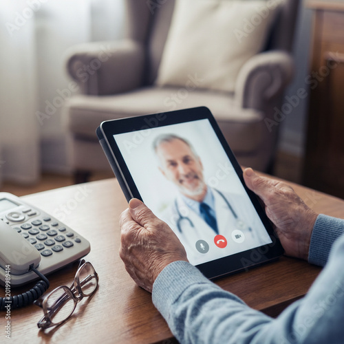 Elderly person having a remote video consultation with a male doctor on a digital tablet at home for telehealth services.